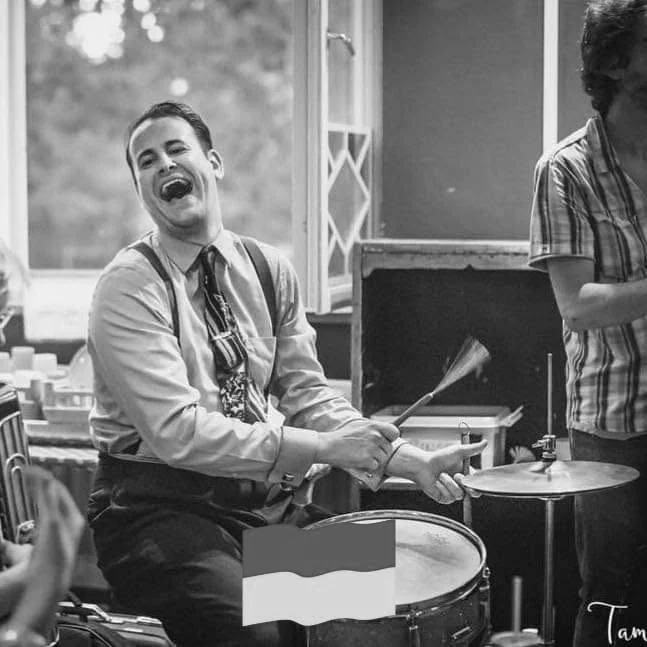 Jon Tigert smiling at the drum kit, playing with brushes.